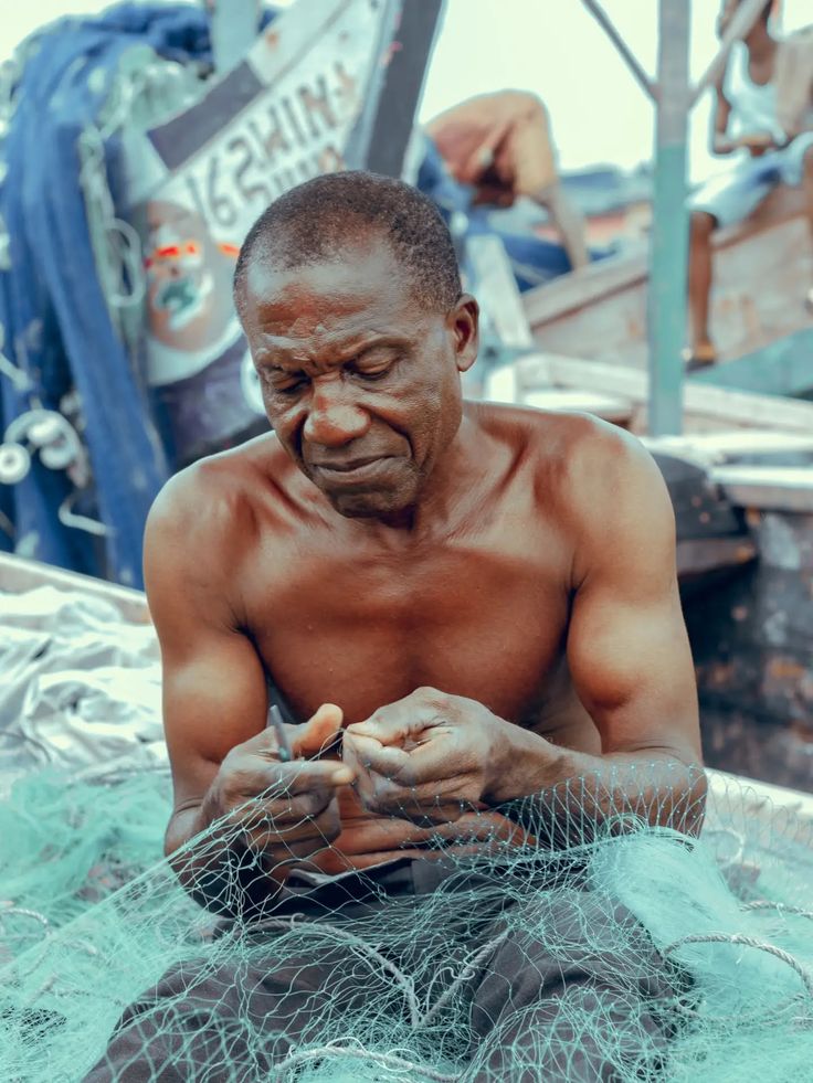 A fisherman mending a green fishing net on a pirogue