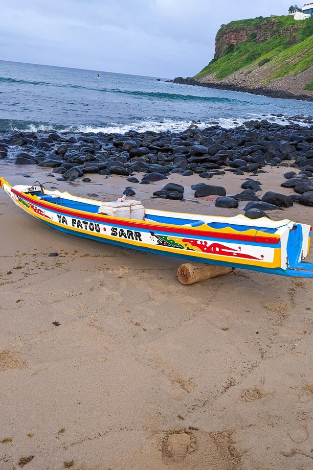 A painted pirogue drawn up on the sand beside black rocks and the sea