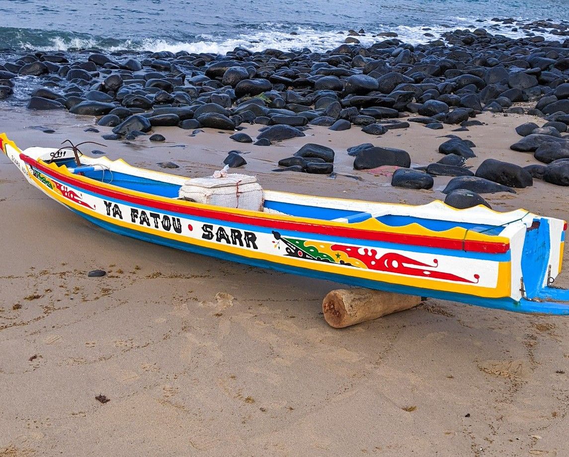 A painted wooden pirogue resting on the sand of a Senegalese beach, below dark rocks at the water&rsquo;s edge